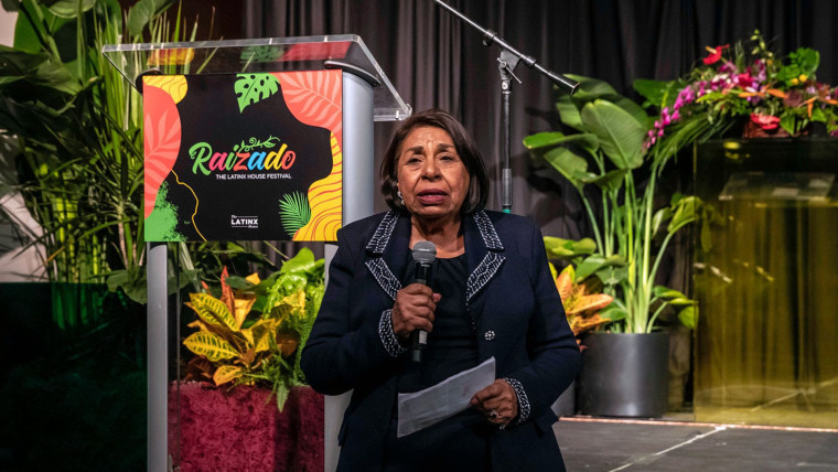 Sylvia Méndez, mujer latina de edad avanzada con un saco azul marino, habla con un micrófono frente a un podio lleno de plantas tropicales