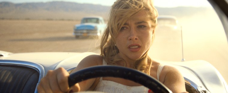 A tight shot on Pugh as she drives a convertible in a desert-like scene. She looks serious and her hair is windswept. A blue, old-fashioned car appears to be following her.