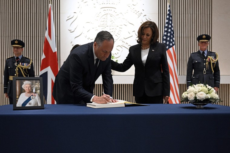 Kamala Harris and Doug Emhoff in black suits sign a book on a table covered in blue fabric. to the left, there is a photograph of the late queen elizabeth. they are in front of both a british flag and an american one.