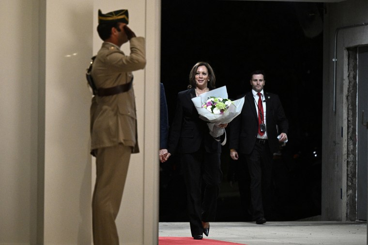 Harris walks toward the camera in a black suit and heels smiles as she carries a bouquet of flowers into a building. A security guard trails behind her and a British officer salutes her as she walks by.