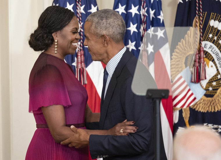 Barack And Michelle Obama hiug in front of the US flag. She is in a pleated pink ombre dress and her hair is in micro-braids and pulled into an elegant bun at the nape of her neck.