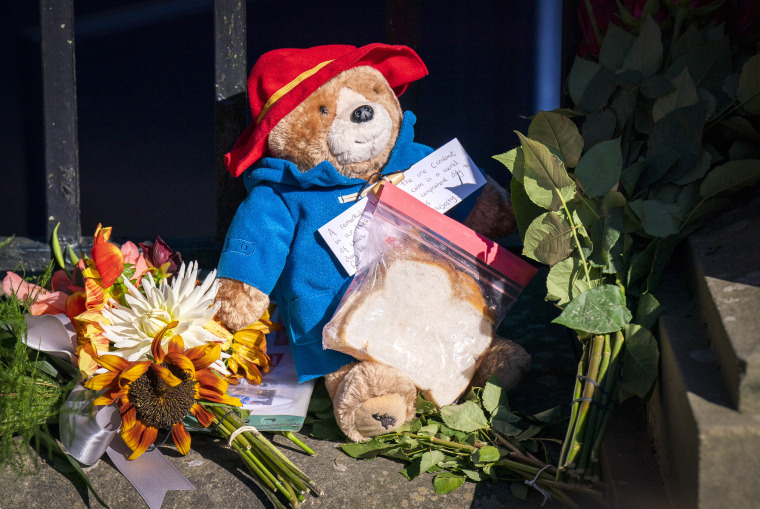 A stuffed paddington bear with a sandwich sits in front of a black gate among flowers.