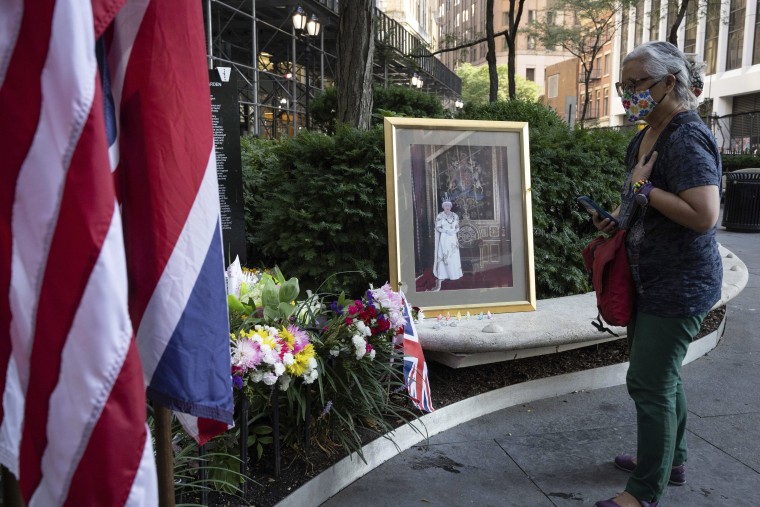 A woman in a T-Shirt, green pants and a red crossbody bag and facemask holds her hand over her heart looking at flowers and a portrait of Queen Elizabeth II in New York City.