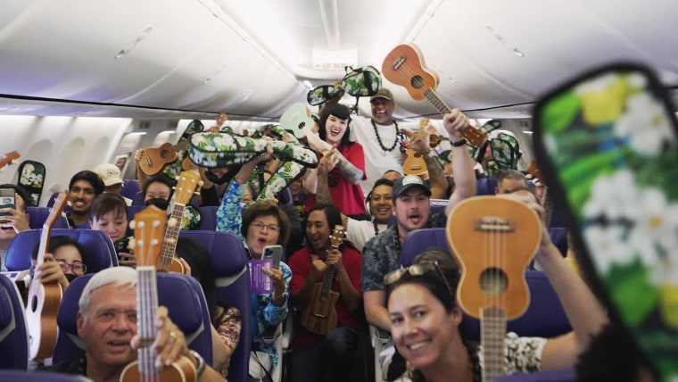 People on a plane hold their ukuleles up in the air, smiling for a camera.