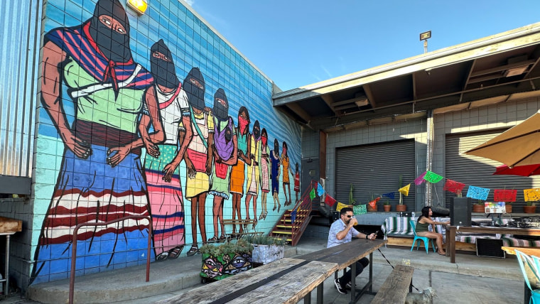 Un hombre en una banca bebe una cerveza mientras revisa su celular. Está sentado frente a un mural de mujeres zapatistas en el restaurante Mujeres Brew House de San Diego