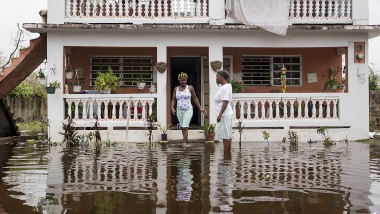 Dos mujeres afroboricuas, con aguas de inundación pasando los tobillos, revisan los daños afuera de una casa en Loíza, Puerto Rico, en septiembre de 2017