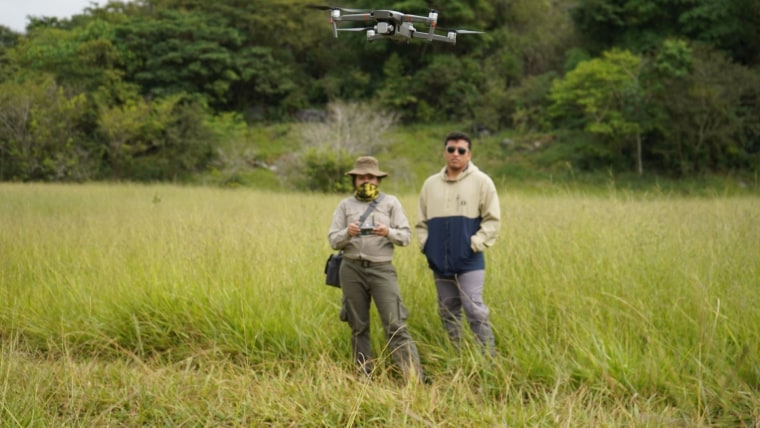 Una mujer con vestimenta de guardabosques y un joven con lentes oscuros usan un control para manejar un dron sobre terreno tropical como parte de una práctica para usar tecnología en el combate contra la deforestación en la Amazonía, en julio de 2022