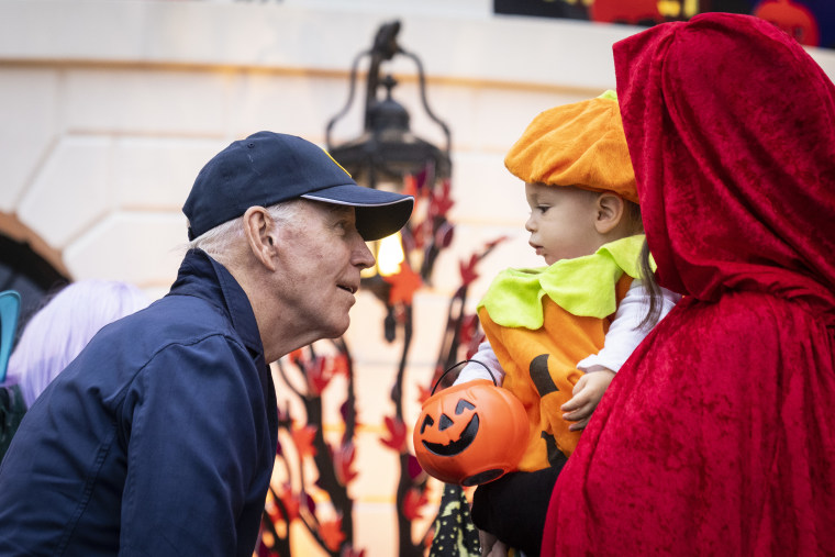 Biden looks at a small child in a pumpkin costume being held by a woman dressed as little red riding hood.