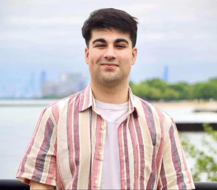 A young man with dark brown hair smiles in a portrait shot in front of the Chicago skyline. HE's wearing a white T-shirt under a red and orange striped button down.