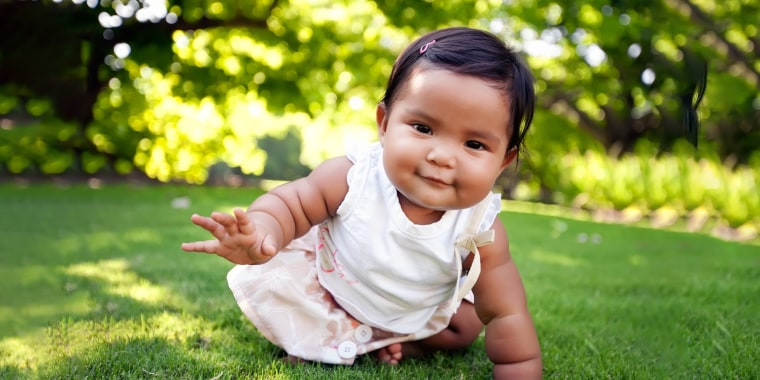 Cute baby girl with a smile on her face, reaching out to take her first crawling step on a lush green lawn at an outdoor park, of mixed ethnic race.