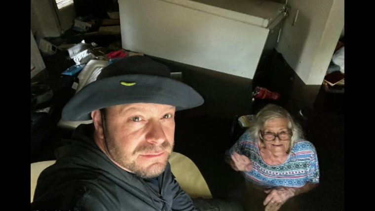 A middle-aged white man in a  gray tilly hat and gray sweater holds a camera aloft for a seflie. His elderly mother with shoulder length white hair smiles, despite being in waist deep brown water inside her home.