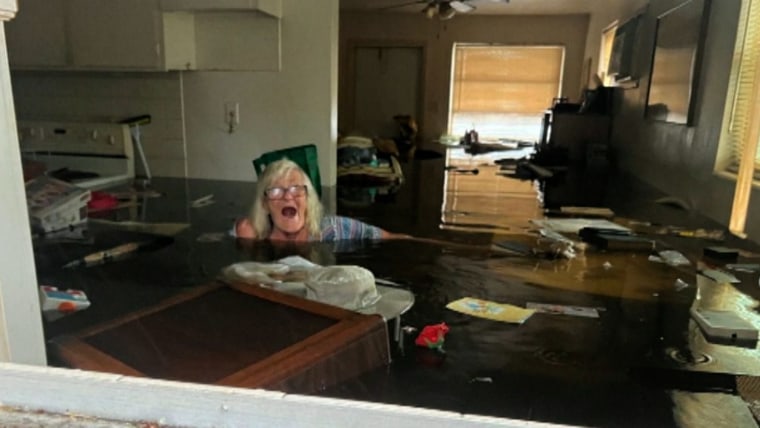An older woman in glasses and a striped shirt stands in neck-deep water in her home surrounded by floating objects. The water appears to be a dark brown.