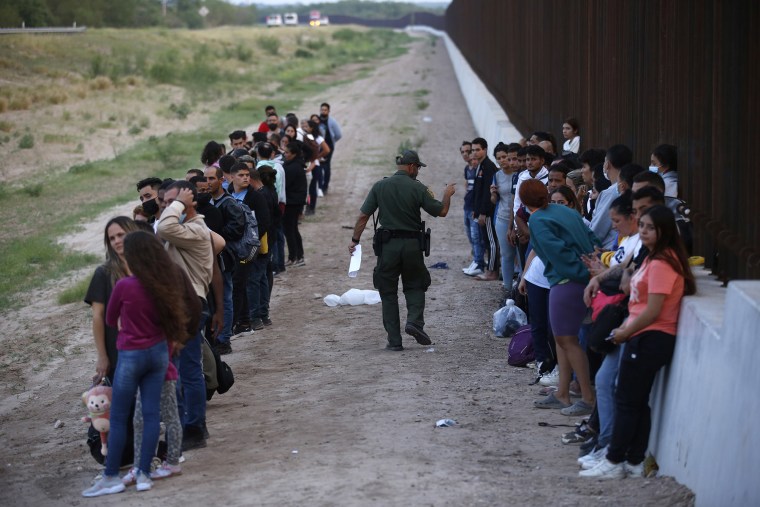 Un grupo de migrantes espera junto al muro fronterizo mientras un agente de la Patrulla Fronteriza hace un recuento en Eagle Pass, Texas, el 21 de mayo de 2022.