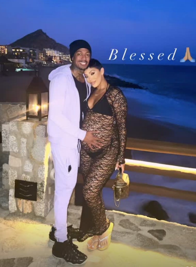 nick cannon and abby de la rosa stand in front of a body of water on a stone walkway with a skyline in the background.