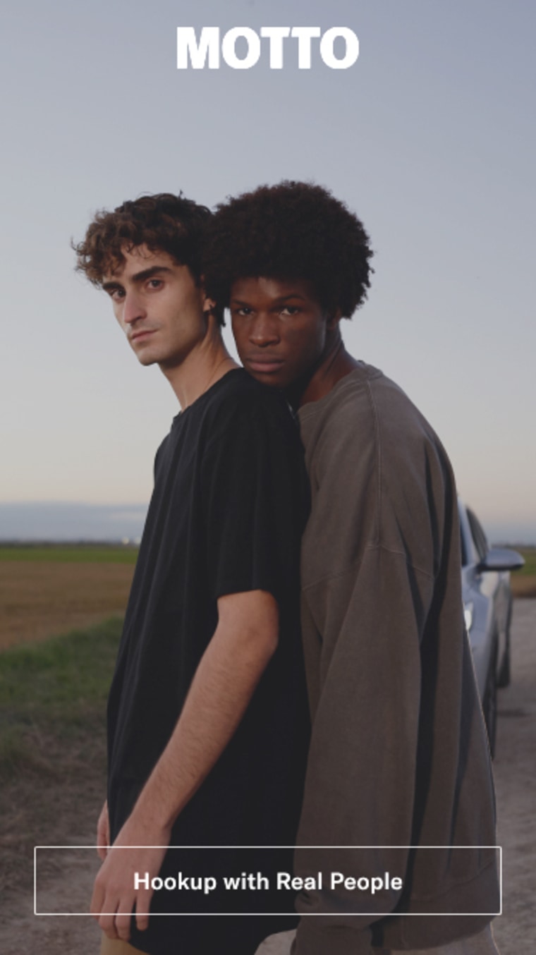 A gay couple standing under the Motto logo.