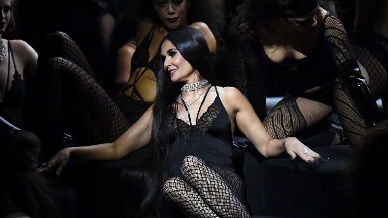 Demi Moore seated onstage, surrounded by backup dancers.