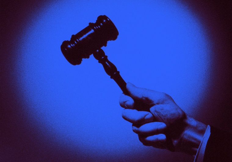 Image: A close up of a judge's hand holding a wooden gavel in a spotlight.