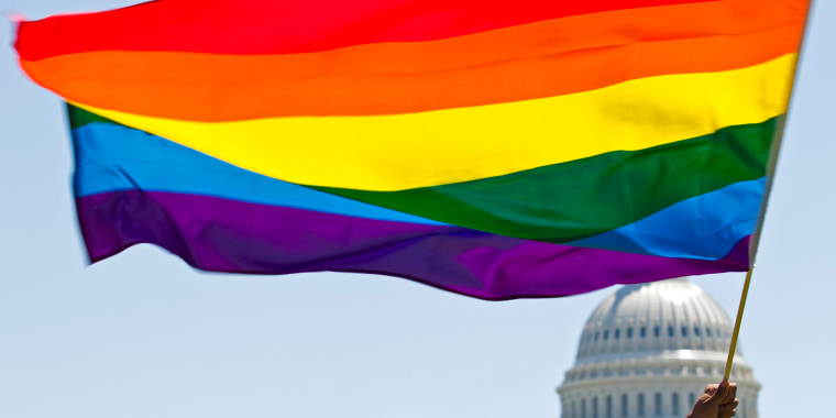 Image: A hand holds up a rainbow flag with the U.S. Capitol dome in the background.