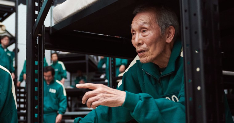 An older man in a numbered green tracksuit sits on a bunk bed, looking off-camera and pointing straight ahead. 