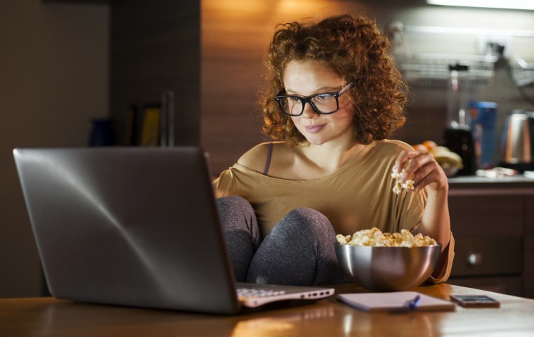Young woman using computer and snacking on popcorn.