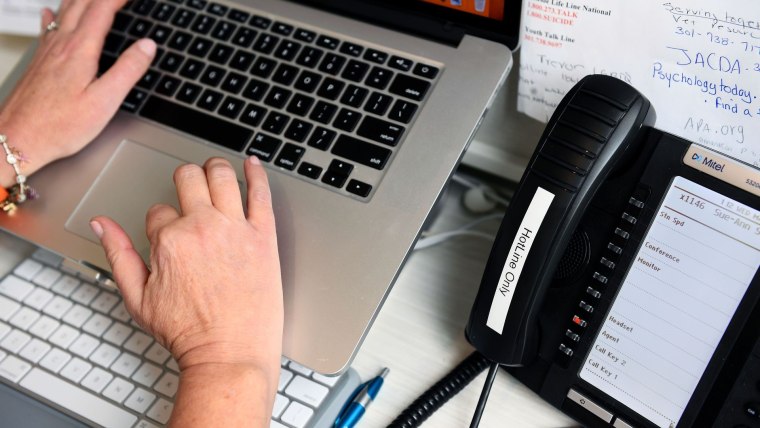 Las manos de una persona están encima de un teclado de computadora Mac al lado de un teléfono que dice "hotline" y es para contestar llamadas de personas teniendo crisis de salud mental