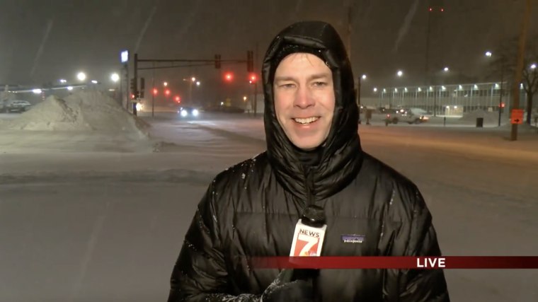 Woodley stands in a black winter coat holding a KWWL microphone outside in the dark with snow falling.