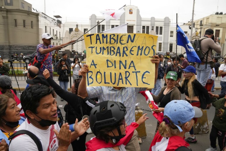 Un grupo de personas peruanas corea y aplaude mientras uno sostiene un cartel que dice "También tumbaremos a Dina Boluarte" en referencia a la primera mujer designada presidenta del país andino.