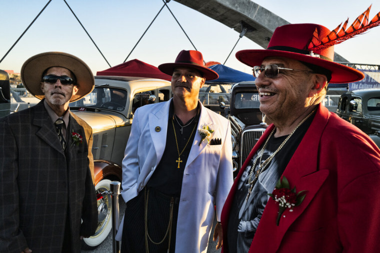 Tres hombres latinos con traje holgado tipo 'zoot suit' y sombreros con plumas clásicos del estilo pachuco posan frente a autos antiguos 