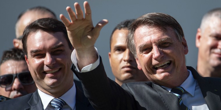 Image: Flavio stands next to Jair Bolsonaro who's waving during a ceremony. 