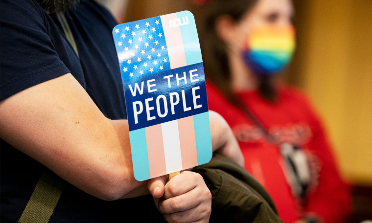 Image: A close up of handing holding a sign that has the transgender pride colors on the American flag with the text,"We the People" and the ACLU logo.