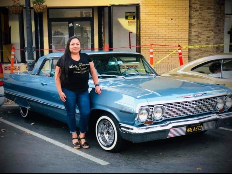 Una mujer latina posa al lado de un auto clásico de color azul cielo que fue arreglado para ser 'lowrider' (tener suspensión cerca del piso). Es integrante del grupo  Lady Lowriders United 