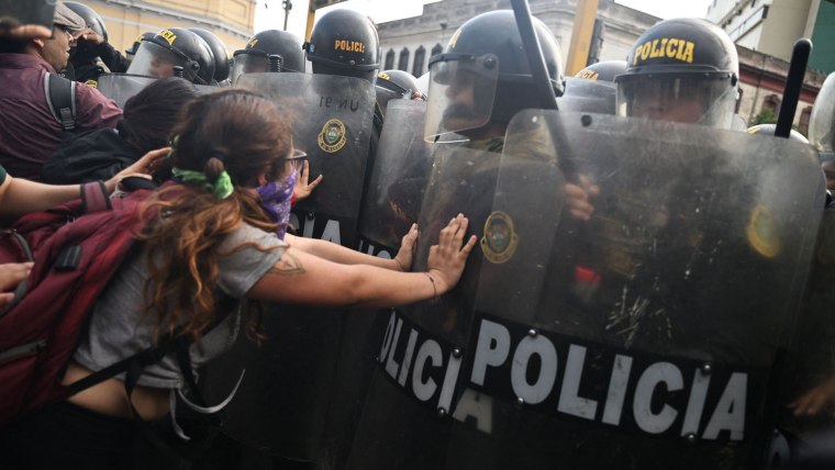 Una joven manifestante de camiseta gris y con un pañuelo morado sobre boca y nariz para protegerse de gases lacrimógenos empuja para atrás a un policía con equipo antimotines. Los policías están formando una fila afuera de un sitio donde estuvieron retenidos más de 200 manifestantes el 21 de enero de 2023