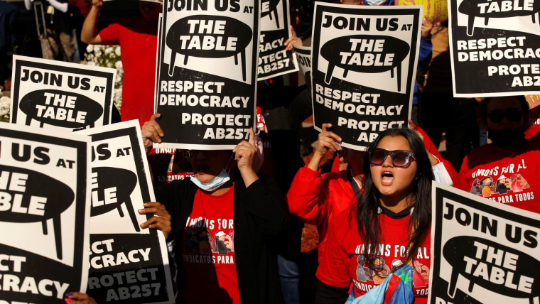 Un grupo de personas protestan en California con camisetas rojas que dicen "sindicatos para todos" y carteles que dicen "Los invitamos a nuestra mesa: respeten la democracia y protejan la ley AB257", que permite a los trabajadores de restaurantes de comida rápida  negociar colectivamente por mejores salarios y condiciones laborales
