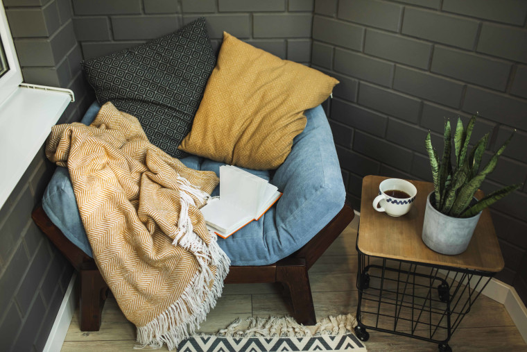 Cozy decorated place for relaxation on balcony. Armchair with pillows and plaid, coffee table, opened book