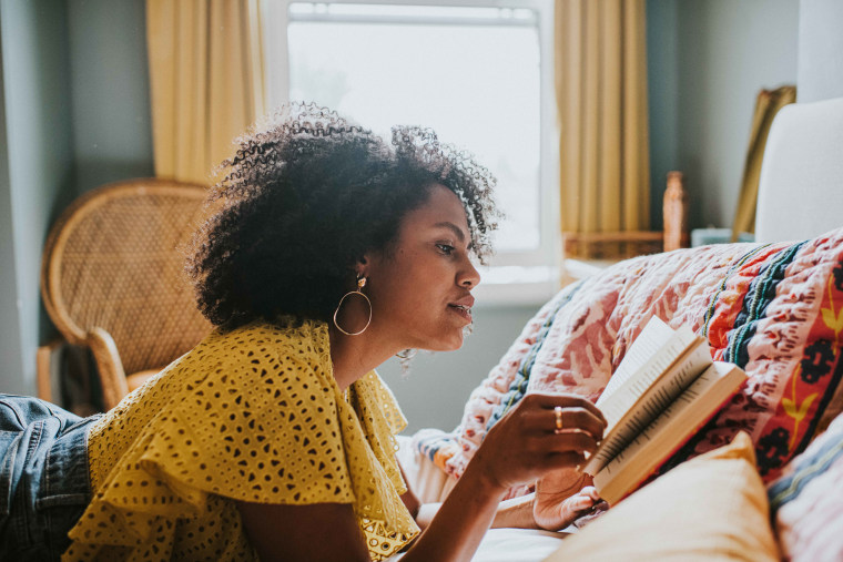 A woman lies on her stomach on a comfortable bed and reads a book