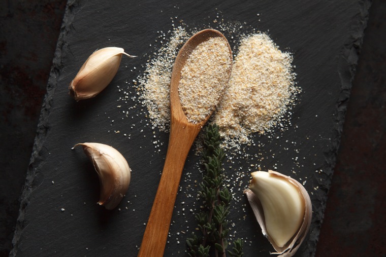 Close up composition of garlic powder sprinkled into wooden spoon, two sprigs of thyme and three cloves on black slate board. Selective focus