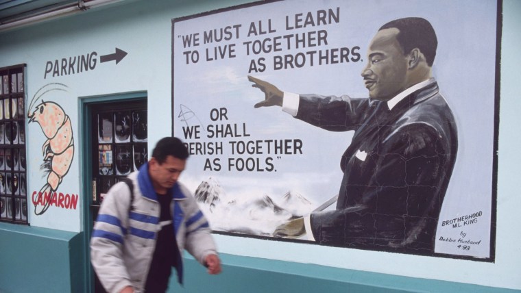 Un hombre latino camina al lado de un mural que muestra a Martin Luther King Jr. con una cita suya qe dice "Debemos aprender a vivir como hermanos o morir juntos como necios". El mural, captado en 1998, está en una zona de Los Ángeles junto a un restaurante de mariscos propiedad de latinos que dice "camarón".