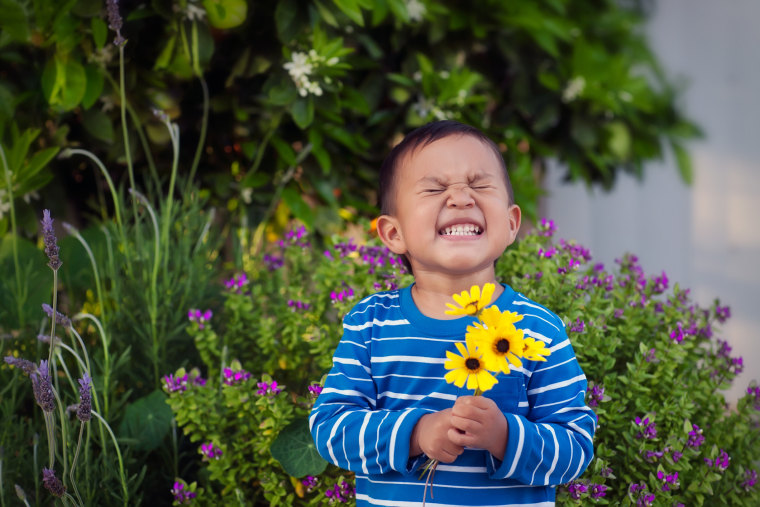 A cheerful young boy holding a few hand picked yellow flowers during spring time, standing in front of a lush garden.