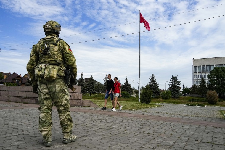 Una joven pareja pasa junto a un soldado ruso que vigila una zona en el Callejón de la Gloria de las hazañas de los héroes el viernes 20 de mayo de 2022, con una réplica de la bandera de la Victoria que marca el 77 aniversario del final de la Segunda Guerra Mundial justo en el fondo. 