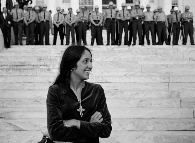Joan Baez, música estadounidense de ascendencia mexicana con un vestido de terciopelo, está parada frente a unas escaleras. En la cima de las escaleras hay una fila larga de muchos policías que pertenecen a la fuerza estatal de Alabama. Es la marcha por los derechos civiles que recorrió desde Selma hasta Montgomery en marzo de 1965