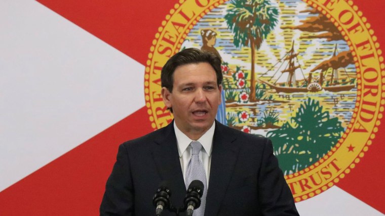 Ron DeSantis habla desde un podio con micrófonos y frente a una pantalla que muestra la bandera estatal de Florida
