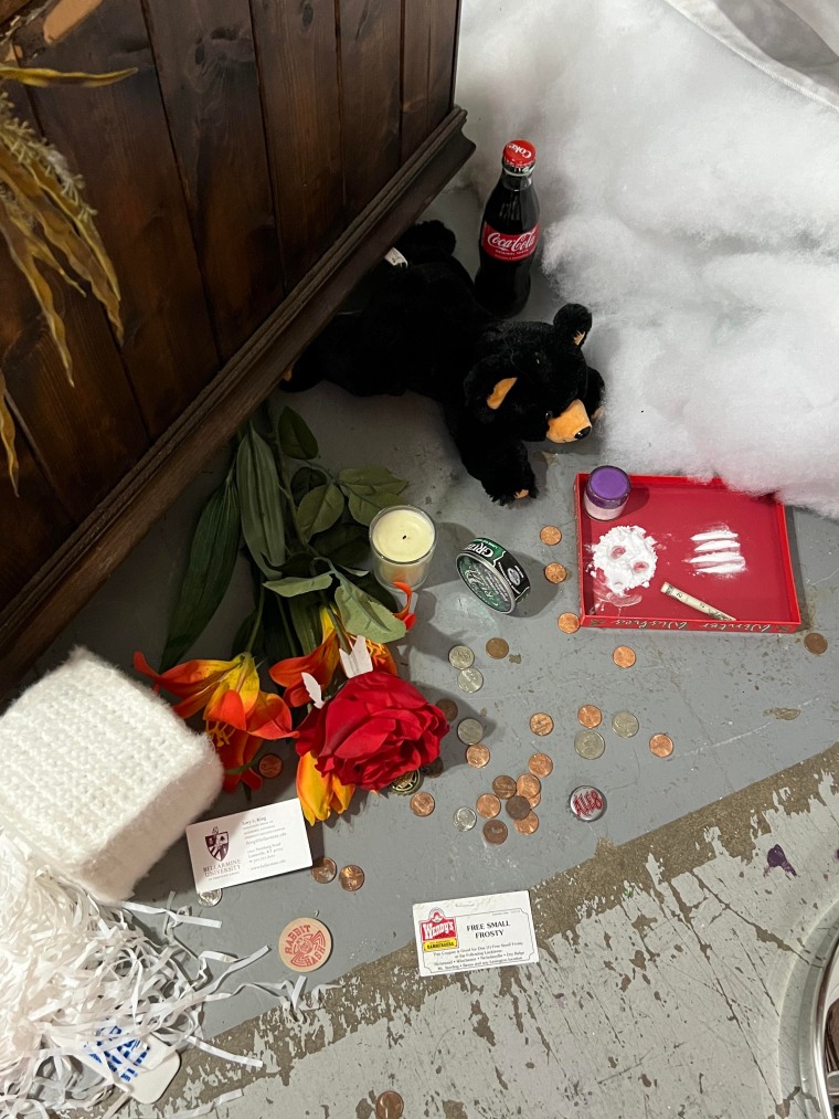 Trinkets — including a stuffed bear, knitted cocaine and fake lines of the drug — line the floor at the base of a taxidermed bear in Kentucky. There's also change, flowers, and a candle.