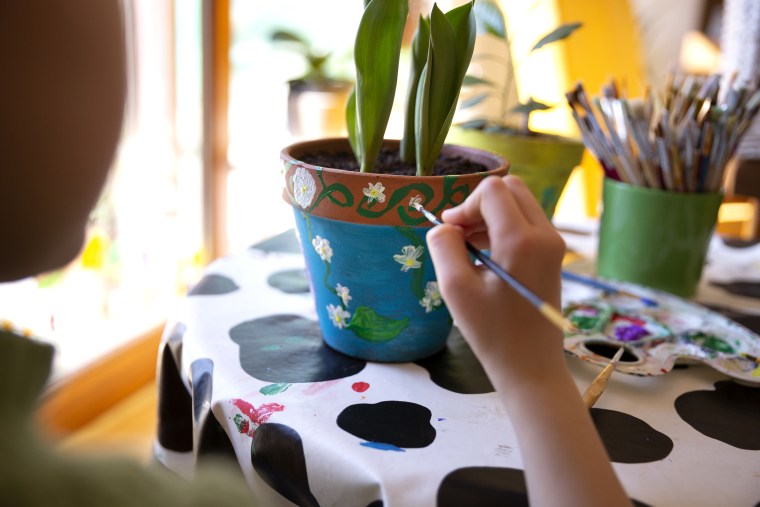 Over the shoulder view of a child painting a terracotta plant pot with bright coloured paints on a dining table in springtime.