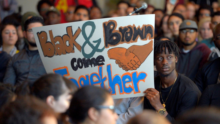 En un auditorio están sentados muchos jóvenes sobre todo negros y latinos. Al centro, un joven sostiene una pancarta que dice "Black and Brown come together" o "Negros y latinos unidos" como parte de una protesta en la Universidad Estatal de California, campus Long Beach, en marzo de 2016