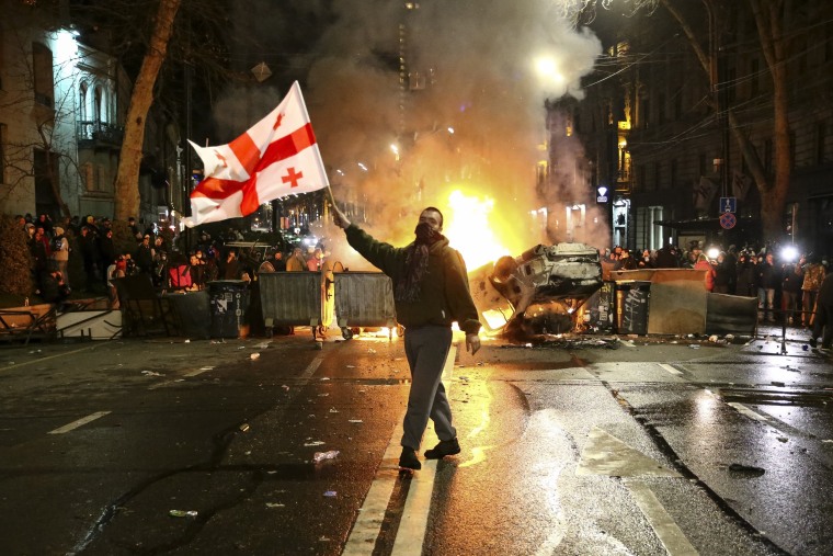 A man waves a Georgian flag in front of a burning barricade as other protesters stand behind not far from the parliament building in Tbilisi on March 9, 2023. 