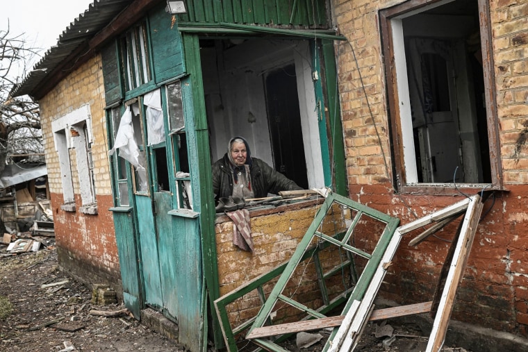 An elderly woman stands behind a window in her damaged house