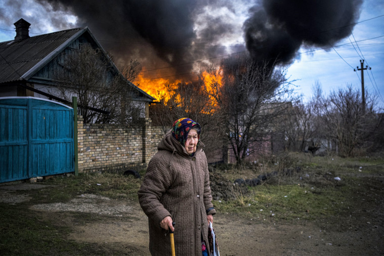 An elderly woman stands near a burning building following shelling