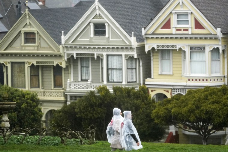 Pedestrians wearing ponchos walk down a path at Alamo Square Park in front of the "Painted Ladies," a row of historical Victorian homes, in San Francisco, Tuesday, March 21, 2023.
