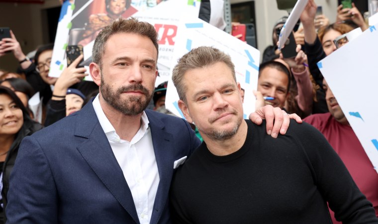 Ben Affleck (left) and Matt Damon stand in front of a cheering crowd with "Air" posters. Affleck is in a suit and collared shirt, Damon is in a black long sleeved crew neck sweater.