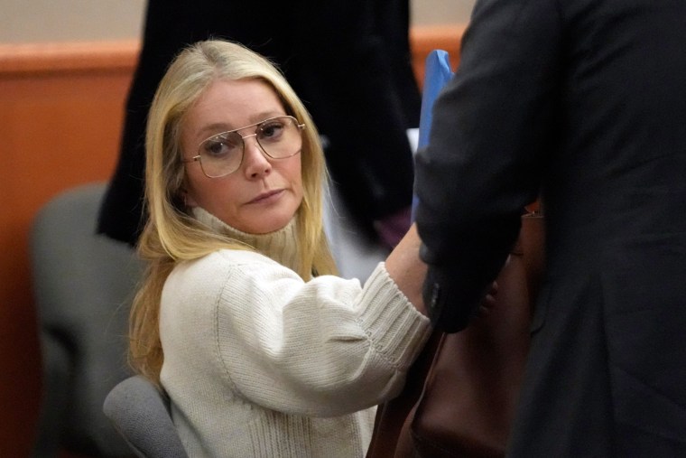 Paltrow in a white turtleneck sweater and aviator-style glasses frowns at something off-screen.